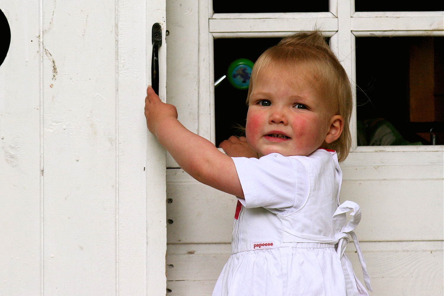 Pretty little blonde girl in a white dress opening the door of a white shed