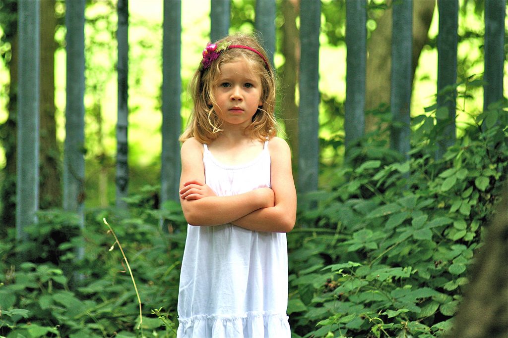 Cute little blonde girl standing with crossed arms in the woods with a scowl on her face