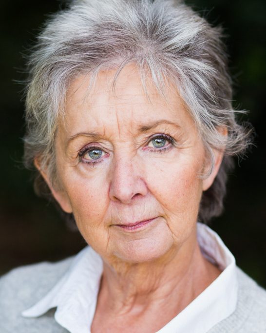 Older actress with grey hair and a white shirt looking into the camera